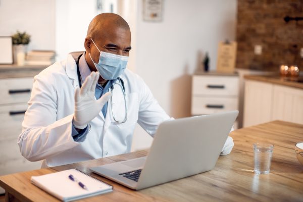 Happy African American doctor using laptop and waving to someone while having video call from his office during coronavirus pandemic.